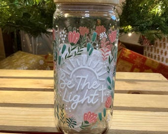May include: A clear glass jar with a wooden lid, decorated with floral patterns in pink, white, and green. The jar features the text "Be The Light" with a cross and sun design. The jar is sitting on a wooden surface.