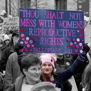 May include: A group of people holding signs at a protest. One sign reads "Thou Shalt Not Mess With Women's Reproductive Rights." The sign is decorated with pink hearts and flowers.