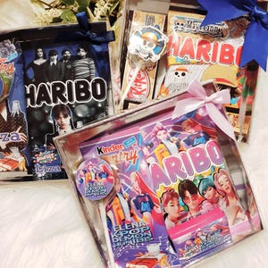 Peut inclure: Deux boîtes cadeaux transparentes contenant des assortiments de bonbons et snacks. Une boîte présente le thème Wednesday Addams avec Haribo et Smarties, nouée d'un ruban bleu foncé. L'autre boîte présente la marque Elena Kpop Demon Hunters avec Haribo, Kinder Country et une sucette, nouée d'un ruban rose clair.