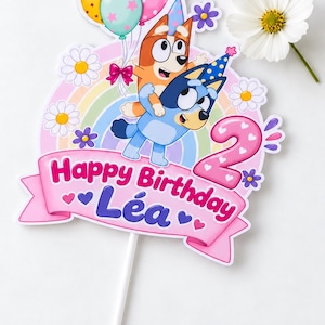 Peut inclure: Une décoration de gâteau représentant les personnages Bluey et Bingo, un arc-en-ciel, des ballons et des marguerites. Une bannière rose affiche le texte Happy Birthday Léa et un grand chiffre 2, le tout fixé sur une tige blanche.
