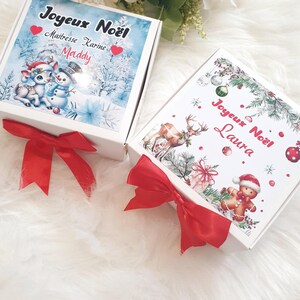 K&ouml;nnte beinhalten: Zwei wei&szlig;e Geschenkboxen mit roten Satinschleifen. Eine Box zeigt eine Winterszene mit einem Schneemann und einem Hirsch, den Text "Joyeux No&euml;l" und den Namen "Maddy". Die andere Box hat einen Lebkuchenmann und den Text "Joyeux No&euml;l Laura".