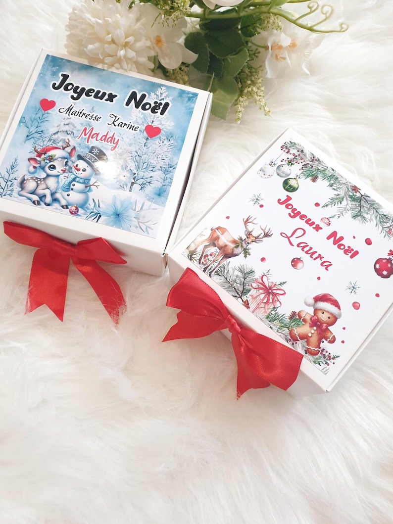 K&ouml;nnte beinhalten: Zwei wei&szlig;e Geschenkboxen mit roten Satinschleifen. Eine Box zeigt eine Winterszene mit einem Schneemann und einem Hirsch, den Text "Joyeux No&euml;l" und den Namen "Maddy". Die andere Box hat einen Lebkuchenmann und den Text "Joyeux No&euml;l Laura".