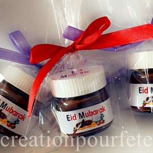 Peut inclure: Trois petits pots en verre de pâte à tartiner au chocolat enveloppés dans du cellophane transparent avec un ruban rouge et un ruban violet. Chaque pot a une étiquette blanche avec le texte "Eid Mubarak" en noir.