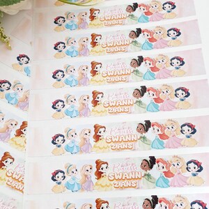 Peut inclure: Étiquettes rectangulaires avec des princesses de dessins animés dans différentes robes. Chaque étiquette porte le texte "Princesse Swann 2 ANS". Les princesses incluent Blanche-Neige, Cendrillon, Belle, etc., dans un schéma de couleurs rose et blanc.