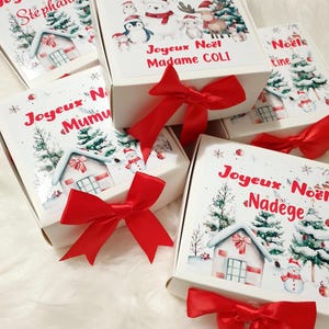 K&ouml;nnte beinhalten: Wei&szlig;e Geschenkboxen mit roten Satinschleifen. Die Boxen sind mit weihnachtlichen Illustrationen wie verschneiten H&auml;usern, B&auml;umen und Schneem&auml;nnern verziert. Auf den Boxen steht "Joyeux No&euml;l" und verschiedene Namen.