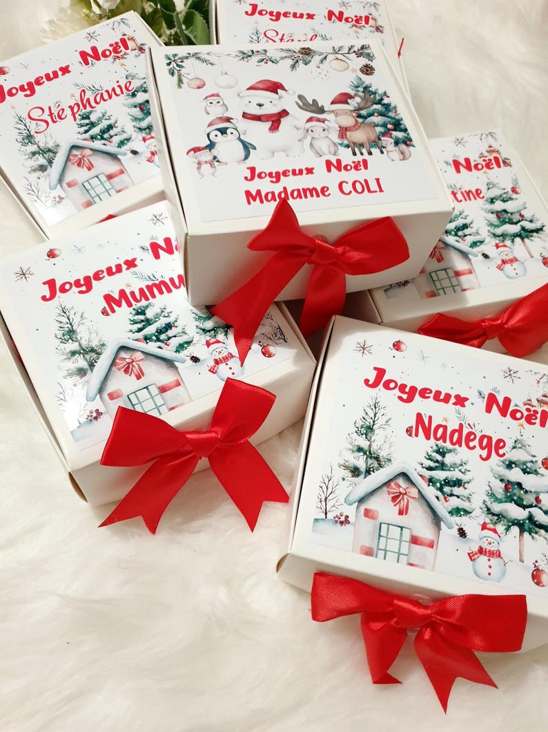K&ouml;nnte beinhalten: Wei&szlig;e Geschenkboxen mit roten Satinschleifen. Die Boxen sind mit weihnachtlichen Illustrationen wie verschneiten H&auml;usern, B&auml;umen und Schneem&auml;nnern verziert. Auf den Boxen steht "Joyeux No&euml;l" und verschiedene Namen.
