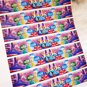 Peut inclure: Sept étiquettes imprimables colorées représentant les personnages du film Vice-Versa. Chaque étiquette a un fond violet, rose et bleu avec une bordure blanche. Le texte "JADE 6 ANS" est imprimé sur chaque étiquette.