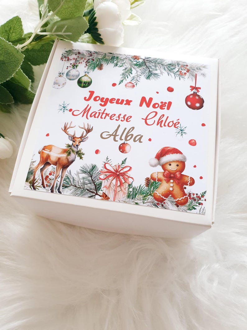 K&ouml;nnte beinhalten: Eine wei&szlig;e Geschenkbox mit weihnachtlichem Design. Die Box zeigt Illustrationen eines Rentiers, eines Lebkuchenmanns, Ornamente und ein verpacktes Geschenk. Auf der Box steht "Joyeux No&euml;l" und "Ma&icirc;tresse Chlo&eacute; Alba."