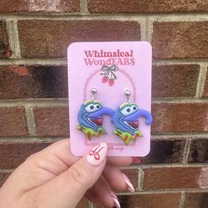 Może przedstawiać: Para niebiesko-zielonych kolczyków z postacią o fantazyjnym wzorze. Kolczyki znajdują się na różowej kartce z tekstem "Whimsical WonDEARS with magic".