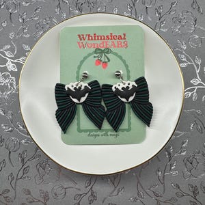 May include: Black and green bow earrings with a bat motif, presented on a green card with the text "Whimsical WondEARS" and "designs with magic". The earrings are on a white plate with a gold rim, against a floral background.