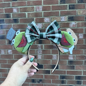 May include: A whimsical headband featuring green frog-themed ears with top hats and bow tie. The ears have red and green accents, and the bow tie is a plaid pattern. The headband is held by a hand against a brick wall.