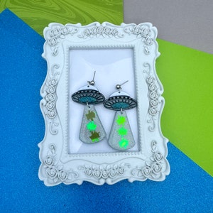 May include: A pair of silver alien-themed earrings. Each earring features a silver UFO design with a teal center and a dangling, glittery, silver teardrop shape with green alien cutouts. The earrings are displayed in a white ornate frame.