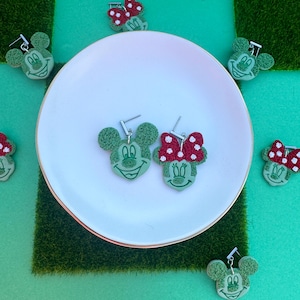 May include: Green Mickey Mouse and Minnie Mouse earrings with silver hooks. The Minnie Mouse earrings have a red and white polka dot bow. The earrings are displayed on a white plate with a gold rim, surrounded by other earrings and a green surface.