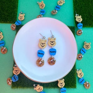 May include: Handmade earrings featuring a smiling bear face, blue "Hunny" pot, and a honey-filled beehive. The earrings are on a white plate, surrounded by similar earrings. The background is green.