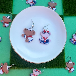 Könnte beinhalten: Modische Ohrringe mit Teddybär-Design in Braun, Rosa, Rot, Weiß und Blau. Die Ohrringe sind auf einem weißen Teller und einer grünen Oberfläche ausgestellt. Die Ohrringe haben silberfarbene Haken.