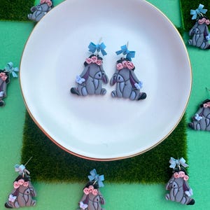 May include: Gray donkey earrings with pink flower crowns and blue bows. The earrings are on a white plate with a gold rim, surrounded by other earrings on a green surface. The donkey earrings have a cartoon-like design.