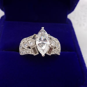 May include: A marquise-cut diamond engagement ring set in a silver band. The ring features a large, clear center stone and smaller accent stones along the band. The ring is displayed in a blue velvet ring box.