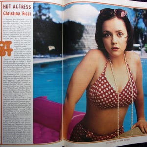 May include: A woman with short dark hair is wearing a red and white polka dot bikini and a pearl necklace. She is sitting on the edge of a swimming pool with her sunglasses on. The photo is from a magazine.