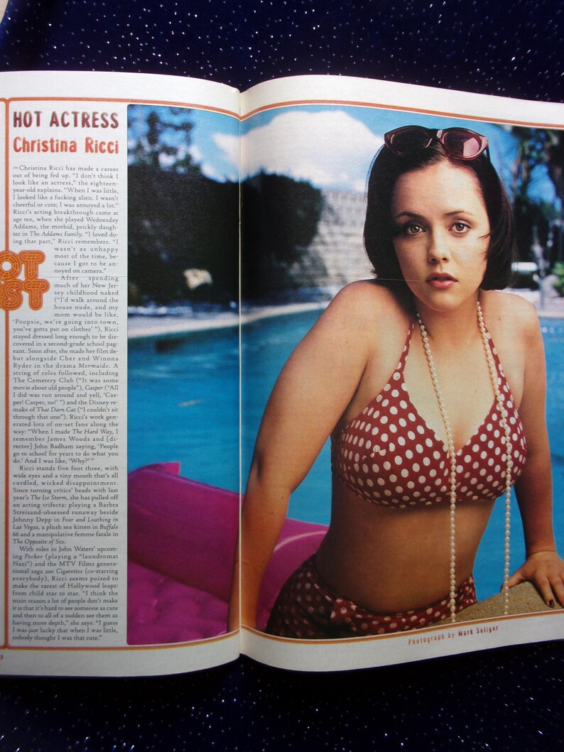May include: A woman with short dark hair is wearing a red and white polka dot bikini and a pearl necklace. She is sitting on the edge of a swimming pool with her sunglasses on. The photo is from a magazine.
