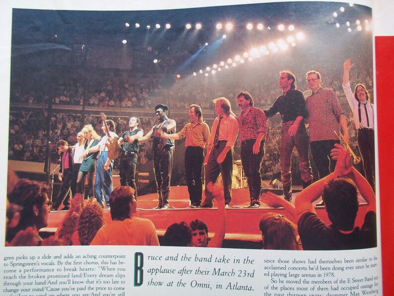 May include: A group of musicians on stage in front of a large crowd. The musicians are taking in the applause after their performance. The text on the page reads "Bruce and the band take in the applause after their March 23rd show at the Omni, in Atlanta."