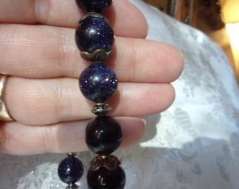 Puede incluir: Primer plano de un collar de cuentas azul oscuro y negro con detalles plateados y bronce. Las cuentas son redondas y tienen una textura moteada.