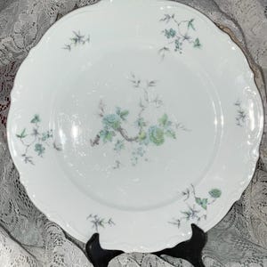 May include: White porcelain plate with a scalloped edge and a floral design in shades of blue and green. The plate features a central branch with stylized flowers and smaller floral accents around the rim. The plate is displayed on a black stand.