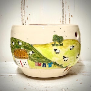 May include: A cream-coloured ceramic yarn bowl featuring a hand-painted pastoral scene of green hills, a white cottage with a thatched roof, and several black and white sheep. A clothesline with colourful laundry hangs near the cottage.