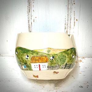 May include: A cream-coloured ceramic yarn bowl featuring a hand-painted landscape scene. The design includes a white cottage with a thatched roof, green rolling hills, black and white sheep, a tree, a stone wall, and small chickens in the foreground.