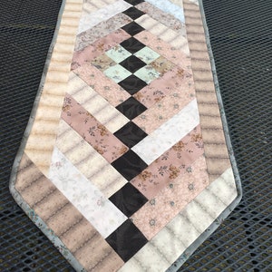 Puede incluir: Camino de mesa acolchado con un diseño de rombos. El camino de mesa presenta telas en tonos beige, marrón, rosa y negro. Los bordes tienen un borde estampado y un ribete gris. Mide unos 120 cm de largo.