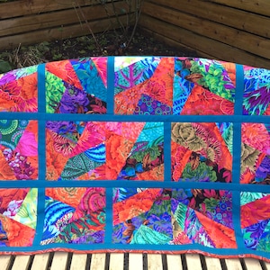 May include: A colorful quilt with a patchwork design featuring various shades of orange, blue, green, and purple. The quilt is made up of squares and rectangles, each with a different pattern. The quilt is bordered with a teal blue fabric.