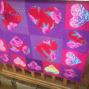 May include: A purple and pink quilt with a heart pattern. The hearts are made of different fabrics with floral prints and sequins. The quilt is hanging on a wooden railing.