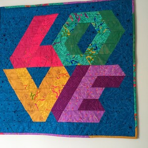 Peut inclure: Une tenture murale matelassée avec le mot "LOVE" écrit en tissu coloré. Les lettres sont composées de différentes formes géométriques et motifs, notamment des hexagones, des triangles et des losanges. Le fond est un tissu bleu foncé avec un motif d'étoiles blanches.