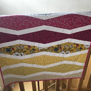 Puede incluir: Una colcha con estampado de chevron en tela rosa, blanca y amarilla. La colcha está colgada en una barandilla de madera.