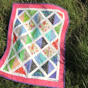 May include: A colorful quilt with a geometric pattern of triangles. The quilt is made of various fabrics with different patterns, including floral, polka dot, and geometric designs. The quilt is bordered with a pink and white trim.