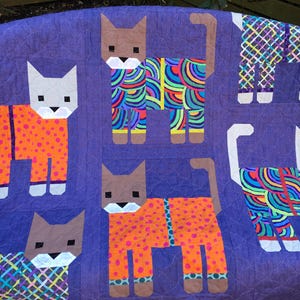 May include: A purple quilt with a patchwork design featuring multiple cats wearing pajamas. The cats are in various poses and have different patterns on their pajamas.