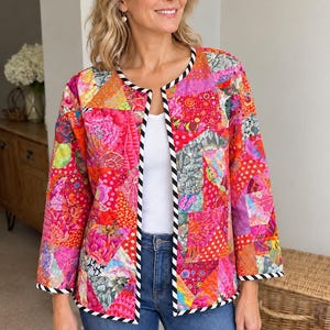 May include: A long-sleeved, open-front jacket featuring a vibrant patchwork design in various shades of pink, orange, and red. The garment is finished with a black and white diagonal striped trim along the collar, front opening, and cuffs.
