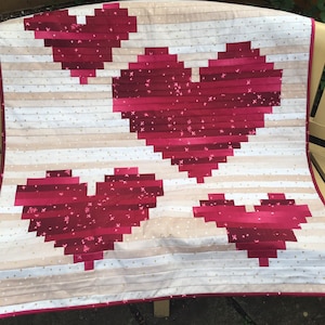 Peut inclure: Une courtepointe avec un fond blanc et trois grands cœurs rouges faits de bandes de tissu. Les cœurs sont disposés en quinconce. La courtepointe est sur un banc en bois.