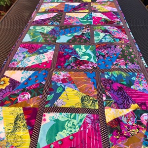 Könnte beinhalten: Ein farbenfroher Patchwork-Tischläufer mit geometrischem Muster. Der Läufer besteht aus verschiedenen Blumenstoffen in den Farben Pink, Lila, Blau, Grün und Gelb. Der Läufer ist mit einem braunen Besatz eingefasst.