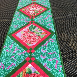Puede incluir: Un camino de mesa verde y rosa con un patrón floral. El camino de mesa está hecho de tela y tiene un diseño acolchado.