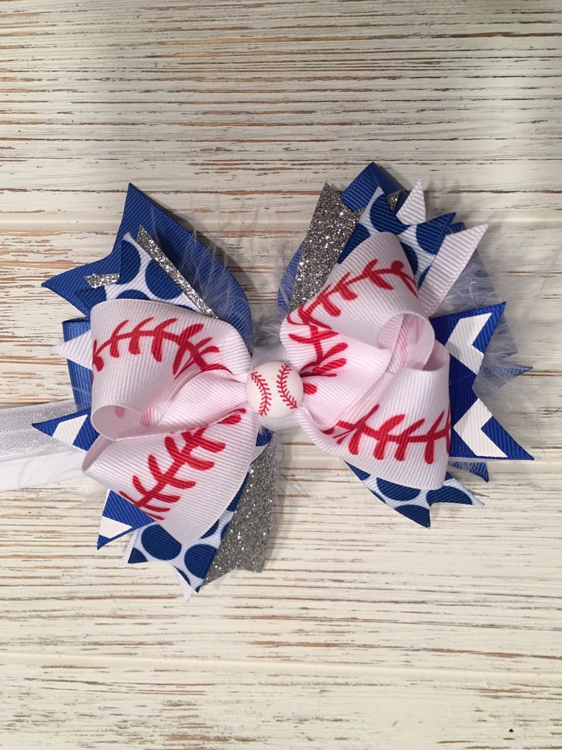 May include: A white, blue, and silver hair bow with a baseball design. The bow has red stitching and a white baseball charm in the center.