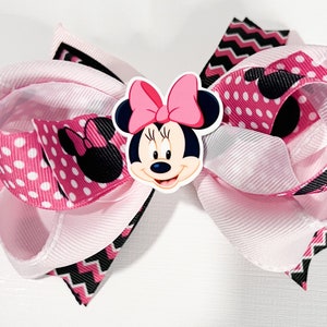 May include: Pink and black bow with a Minnie Mouse character in the center. The bow is made of polka dot and chevron patterned ribbon.