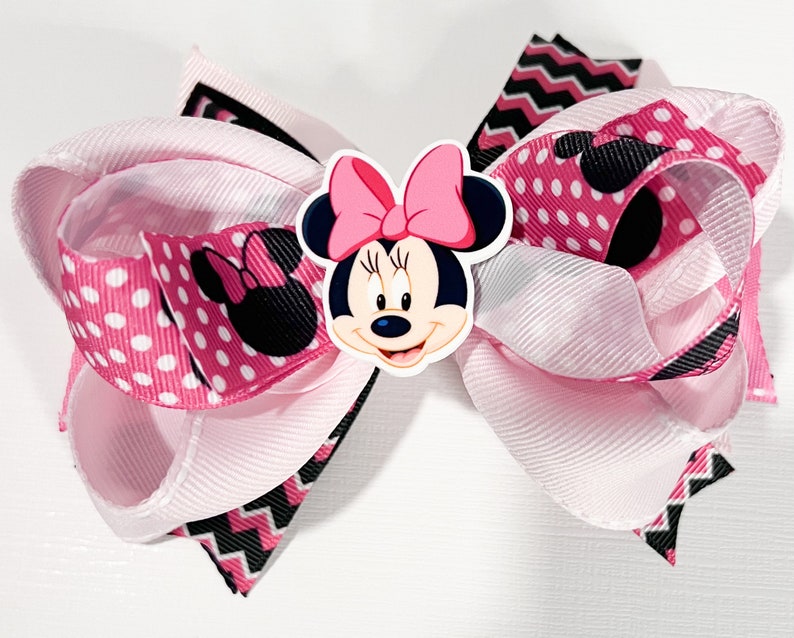 May include: Pink and black bow with a Minnie Mouse character in the center. The bow is made of polka dot and chevron patterned ribbon.