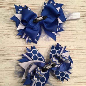 May include: Two blue and white hair bows with a small toy police car charm. The bows are made with blue and white polka dot ribbon, blue and white chevron ribbon, and blue and white striped ribbon.