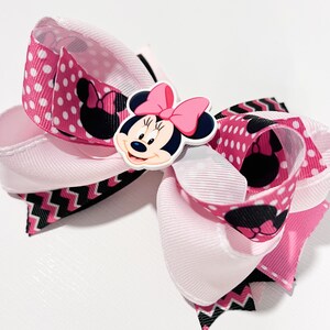 May include: A pink and black bow with a white center and a Minnie Mouse character in the center. The bow is made of ribbon with polka dots and a chevron pattern.