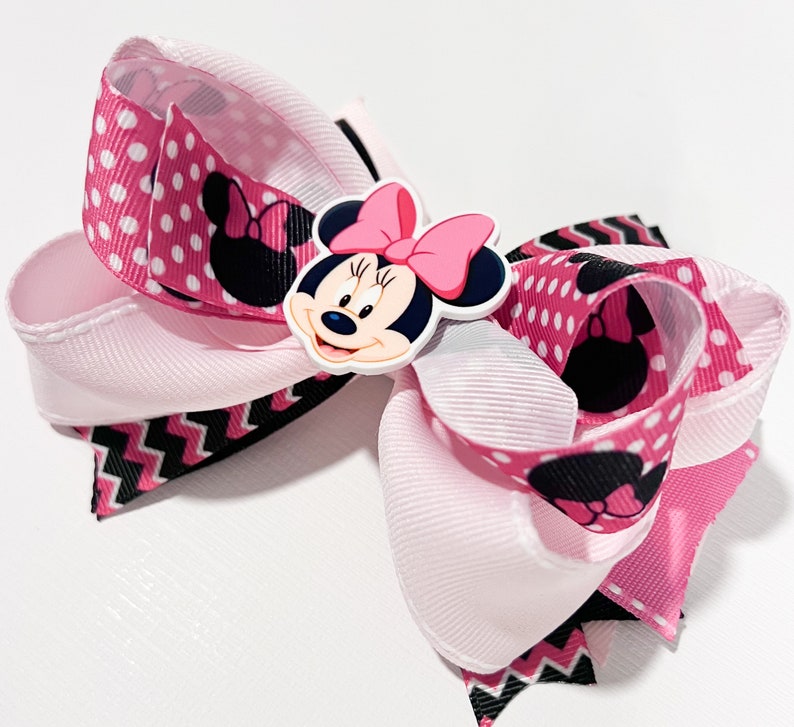 May include: A pink and black bow with a white center and a Minnie Mouse character in the center. The bow is made of ribbon with polka dots and a chevron pattern.