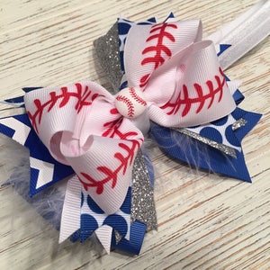 May include: A white, blue, and silver hair bow with a baseball-themed design. The bow features red stitching, white polka dots, and a silver glitter accent. A small white baseball with red stitching is in the center of the bow.