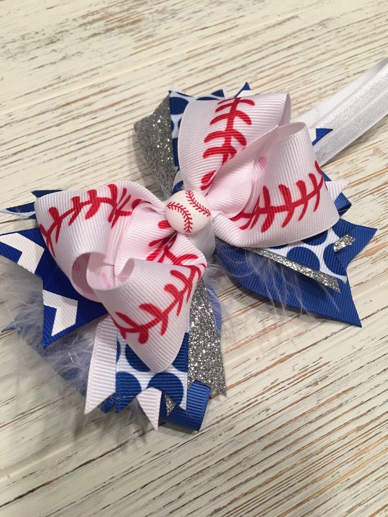 May include: A white, blue, and silver hair bow with a baseball-themed design. The bow features red stitching, white polka dots, and a silver glitter accent. A small white baseball with red stitching is in the center of the bow.