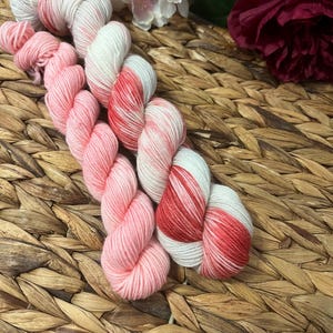 Op de afbeelding: Verschillende strengen garen in roze, witte en rode tinten zijn gerangschikt op een geweven oppervlak. Het garen is zacht en pluizig, met een gevarieerd kleurenpatroon. Roze en witte bloemen zijn op de achtergrond te zien.