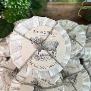 Könnte beinhalten: Ein Stapel runder, weißer, papierverpackter Seifen mit einer braunen Kordelschleife. Jede Seife hat ein Etikett mit einer Schwarzweißillustration eines Schafes und dem Text "Savon & Sippe" und "Savon pour laine".