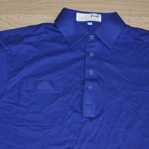 May include: A blue polo shirt with a single chest pocket and a button-up collar. The shirt has a white label with the text "Walter Pringle" on it.